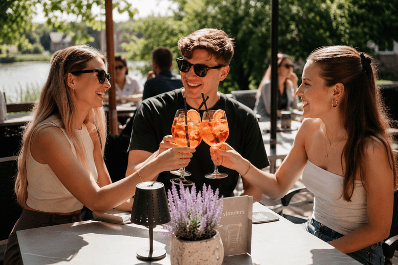 3 personen zitten op het terras aan het water in Utrecht en proosten met een Aperol Spritz